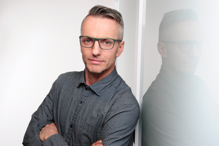 Ein älterer Mann in einem grauen Anzug und einer Brille steht vor einem Bücherregal mit vielen Büchern und einer Weltkugel.