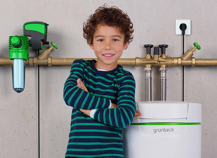 Das Bild zeigt einen Jungen mit lockigem, braunem Haar, der lächelnd vor einer modernen Wasseraufbereitungsanlage steht. Er trägt ein gestreiftes, grünes und blaues T-Shirt und hat die Arme verschränkt. Im Hintergrund sind Rohre und ein Wasserfilter sichtbar, die an einer Betonwand montiert sind.