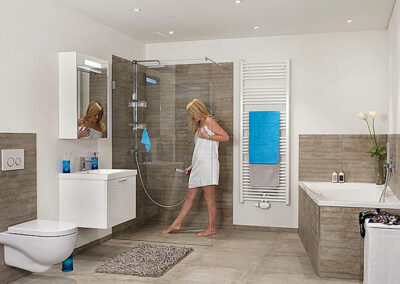 Das Bild zeigt ein modernes Badezimmer mit einer Dusche, einem WC und einer Badewanne. Eine blonde Frau in einem weißen Handtuch steht vor der Dusche. Die Wände sind in neutralen Tönen gehalten, und es gibt einen großen Spiegel sowie einen Handtuchhalter mit blauen Handtüchern. Ein grauer Teppich liegt vor der Dusche.